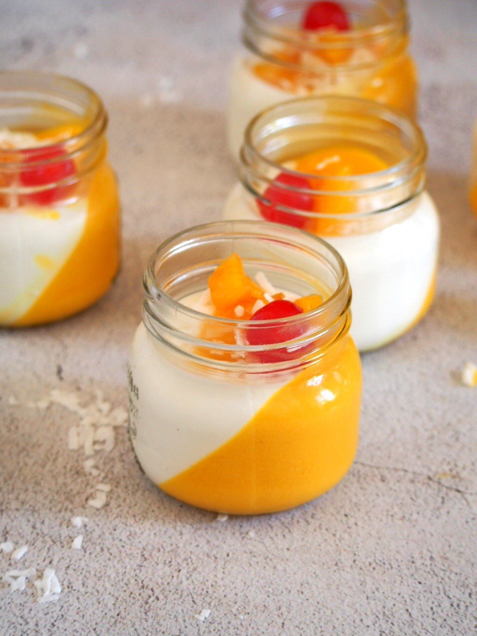 Coconut Mango panna cotta in jars, with cherries, caramelized mangoes and flaked coconut on top.