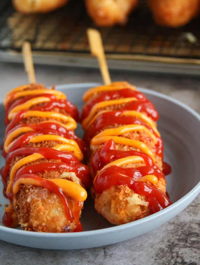 Two Korean Corn Dogs on a plate, drizzled with ketchup and sriracha mayo.