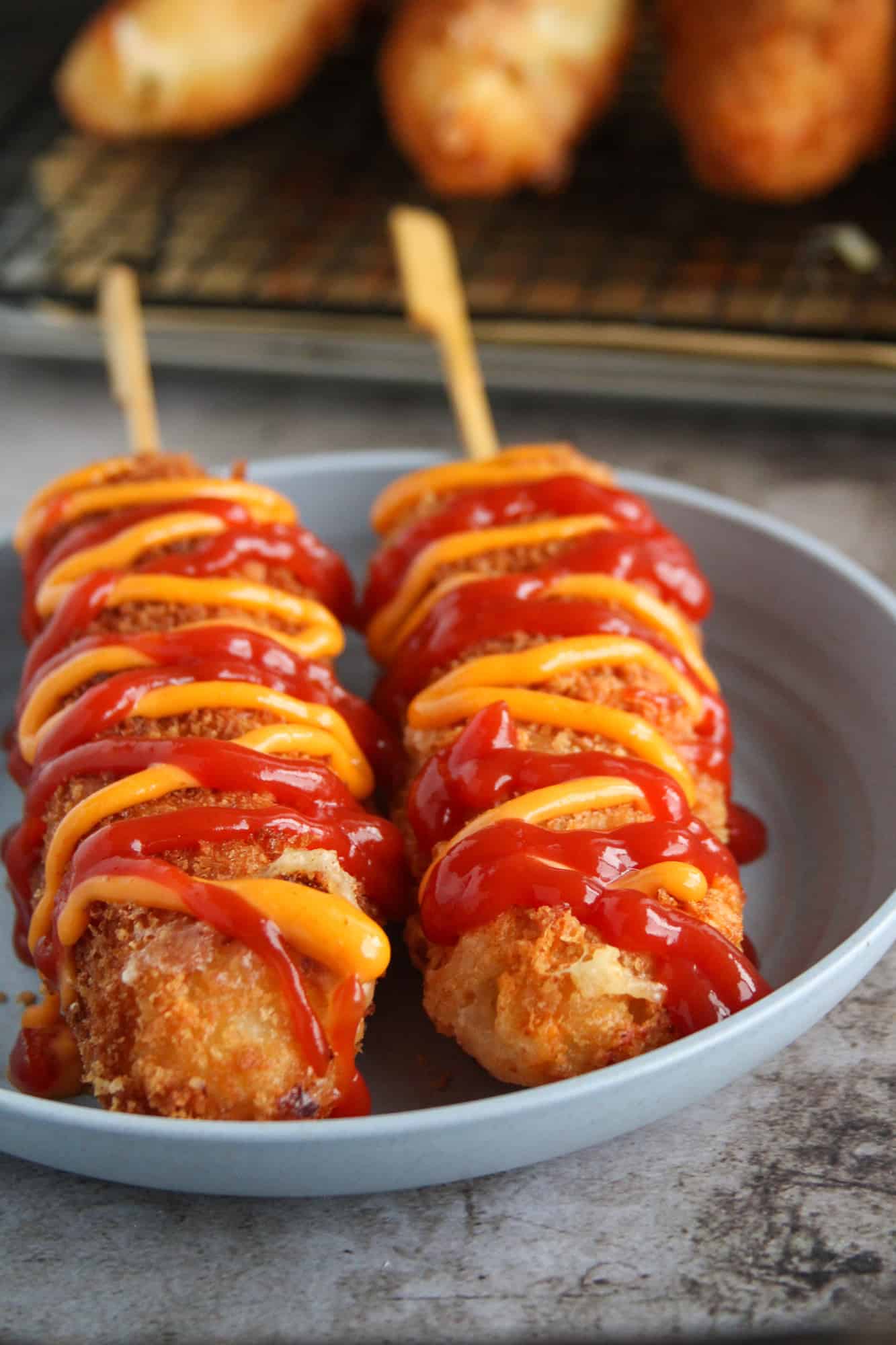 Two Korean Corn Dogs on a plate, drizzled with ketchup and sriracha mayo.