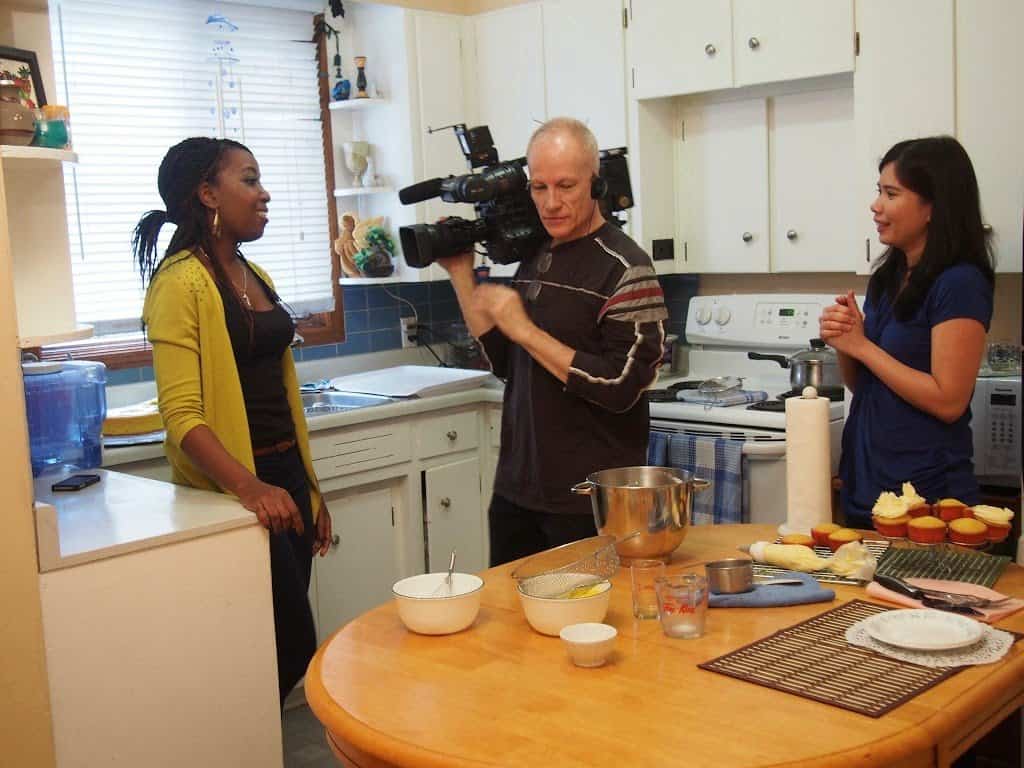 On the set of shoot.  2 ladies with a camera man on a kitchen.